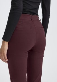 Bordeauxkleurige fitted broek van gladde stof, met achterzakken en een hoge taille, gecombineerd met een zwart langslipshirt.