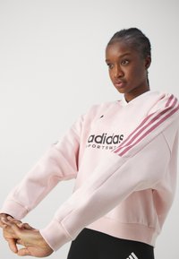 Lichtroze sweatshirt met een ronde hals, zwart Adidas-logo en bordeauxrode drie-streepaccenten langs de mouwen. Zachte stofstructuur.