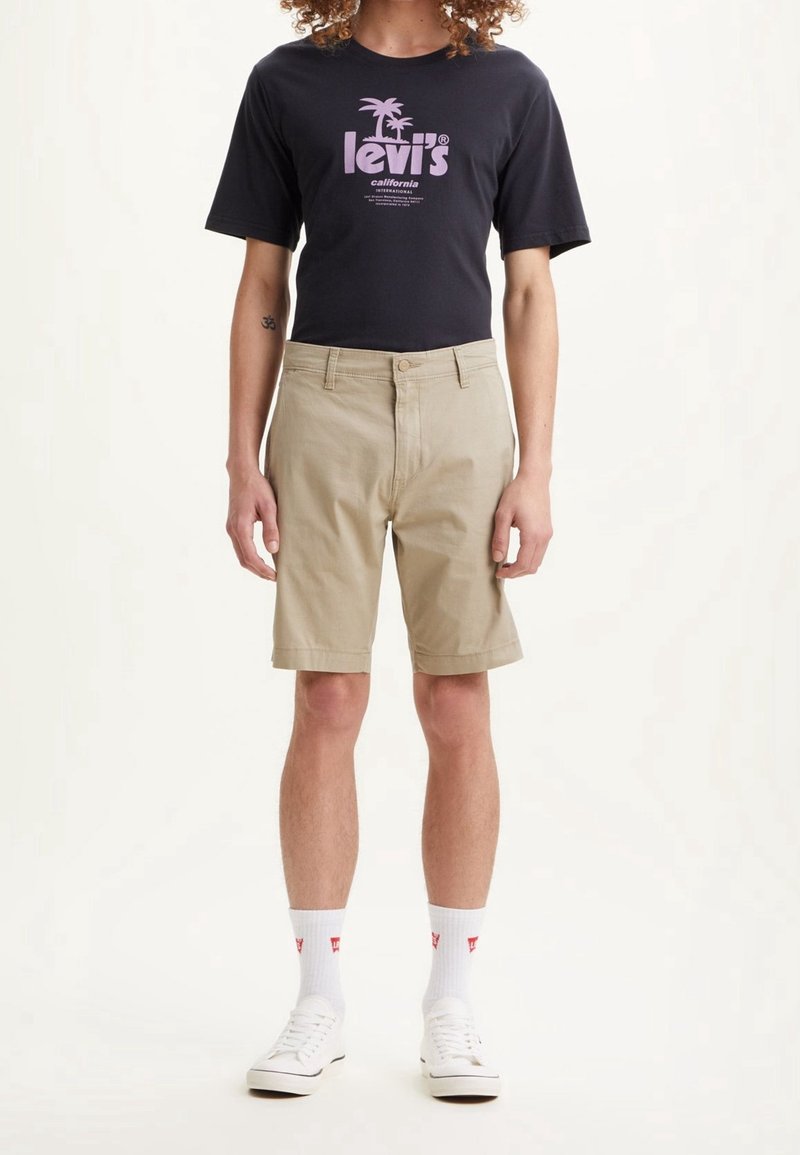 Personne portant un t-shirt noir Levi's, un short beige mi- longueur, des chaussettes blanches avec un logo rouge, et des baskets blanches, debout devant un fond blanc.