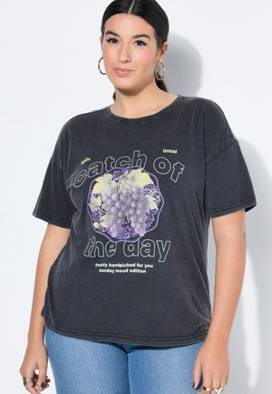 Nainen, jolla musta oversize-t-paita, jossa viinirypälegrafiikka ja teksti "päivän saalis, vastapoimittu juuri sinulle, sunnuntain tunnelma -painos" ja siniset farkut.