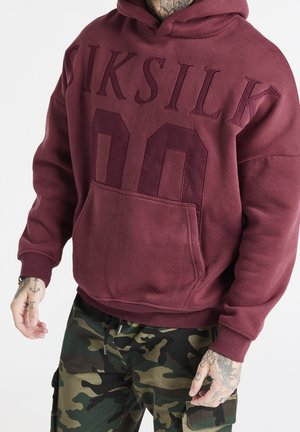 Mężczyzna w bordowym kapturze SikSilk i maskujących spodniach cargo, z widocznymi tatuażami na rękach na białym tle.