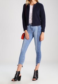 Veste courte texturée bleu marine portée sur un haut blanc, associée à un jean skinny bleu clair à bords effilochés et des bottines noires à talons blocs ouverts à l'avant.