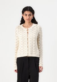 Strikket kremfarget cardigan med diamantmønster, rund hals og knappelukking. Har lange ermer og en kort silhuett.