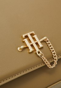 Ręczna torebka z jasnobrązowej skóry, z zamknięciem w postaci metalowego logo w złotym odcieniu oraz ozdobnym łańcuszkiem. Tekstura jest gładka, z widocznym przeszyciem.