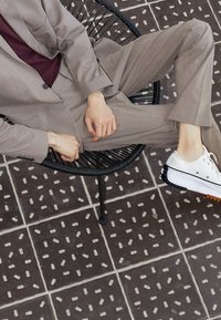 Ljust grå kostym med ficka, bärs över en vinröd skjorta, i kombination med vita sneakers med svart och orange sula, sittande på en svart stol.