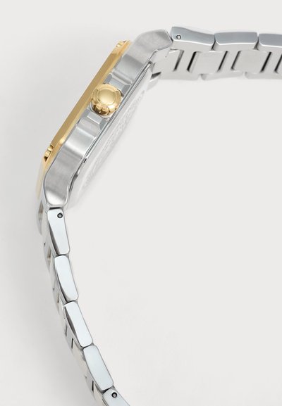 Vue latérale d’une montre en métal bicolore avec une couronne en or et des maillons de bracelet argentés sur fond clair.