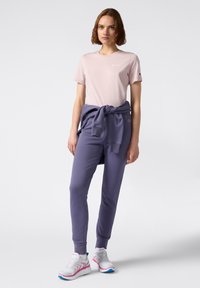 T-shirt di cotone rosa chiaro con un piccolo logo, abbinata a pantaloni della tuta viola scuro con vita annodata. Scarpe da ginnastica bianche e rosa.