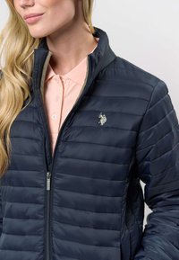 Tamsiai mėlyna izoliuota striukė su siūtais raštais, pilnu užtrauktuku ir mažu išsiuvinėtu logotipu. Dėvima virš švelniai rožinės spalvos marškinių su apykakle.