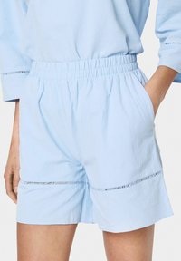 Gaiši zili īsie šorti no mīksta auduma, ar elastīgu jostasvietu, sānu kabatām un kontrastējošu šuvju detaļu gar apakšmalu.