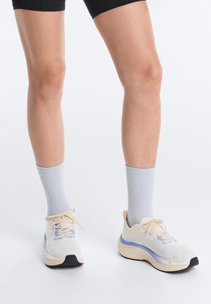 Persoană care poartă șosete gri deschis și pantofi de alergare bej cu accente albastre, stând pe un fundal alb.