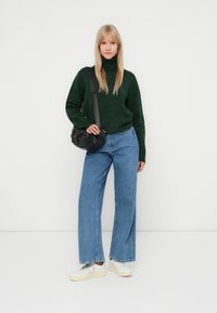 Pull à col roulé vert, jean large bleu clair, sac bandoulière noir et baskets blanches avec des accents gris, sur fond uni.