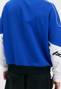 Sudadera azul y blanca con detalles en negro, que cuenta con un dobladillo acanalado y un texto negro distintivo en la manga. Tela suave de peso medio.