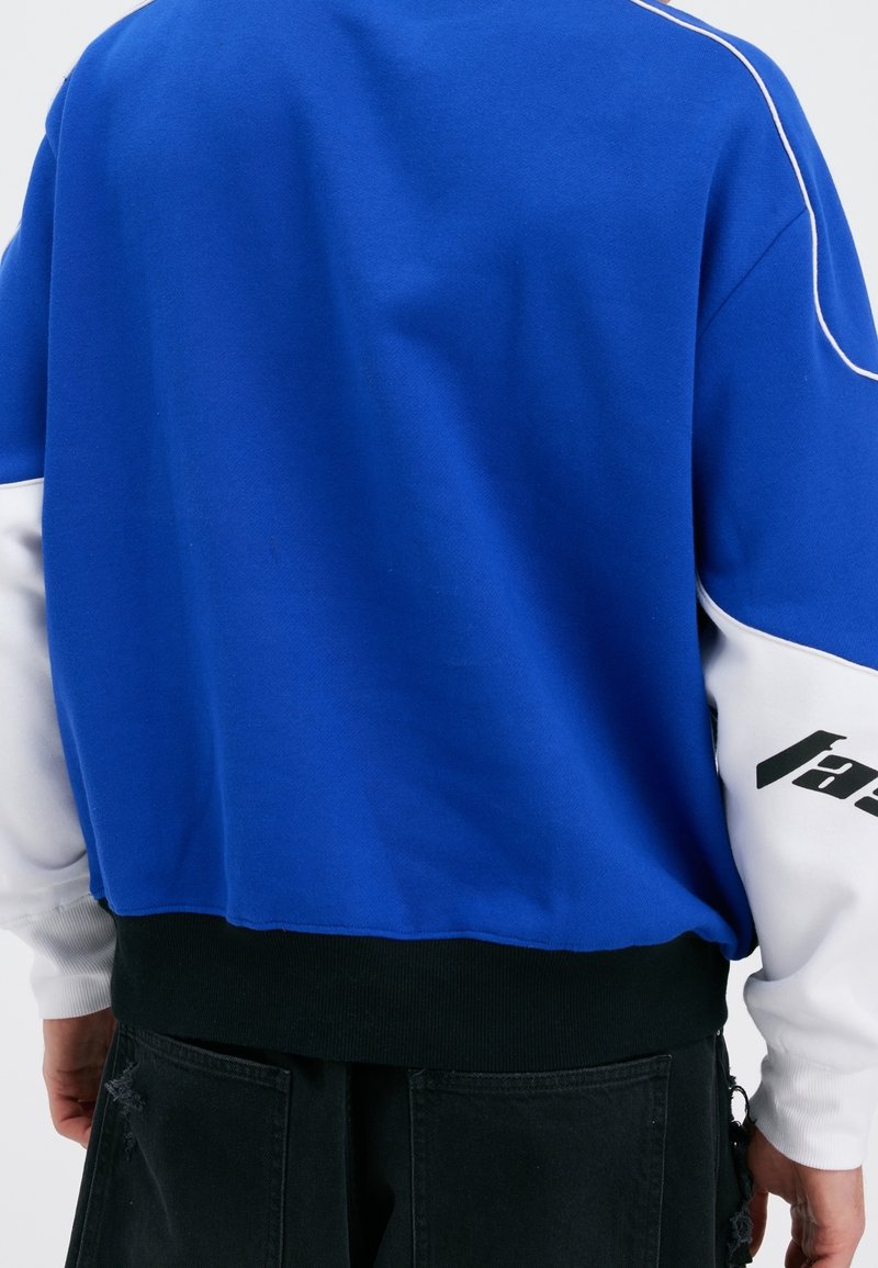 Sudadera azul y blanca con detalles en negro, que cuenta con un dobladillo acanalado y un texto negro distintivo en la manga. Tela suave de peso medio.