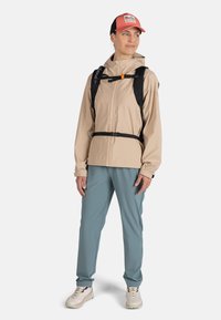 Beige vattentålig jacka med huva, svarta ryggsäcksslingor, teal-byxor och lätta sneakers, kompletterat med en röd keps med en logotyp.