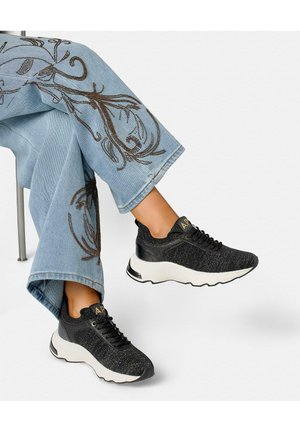 Gekruiste benen met lichtblauwe geborduurde jeans en zwarte chunky sneakers met witte zolen en gouden logo detail op de tong.