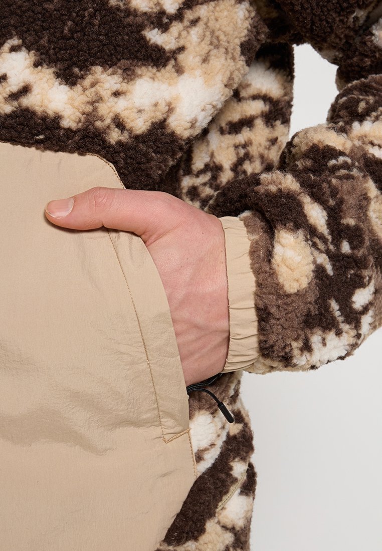 Veste en polaire à motifs marron et crème avec une poche beige, dotée de poignets élastiques et d'un tissu texturé et doux.