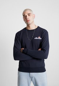 Sudadera azul marino con cuello redondo y puños acanalados. Presenta el logotipo "ellesse" en el pecho. Tejido suave, corte relajado, combinada con jeans claros.