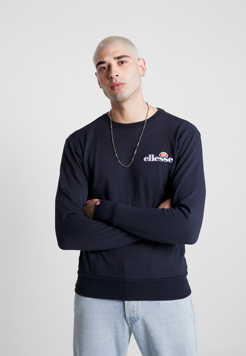 Sudadera azul marino con cuello redondo y puños acanalados. Presenta el logotipo "ellesse" en el pecho. Tejido suave, corte relajado, combinada con jeans claros.