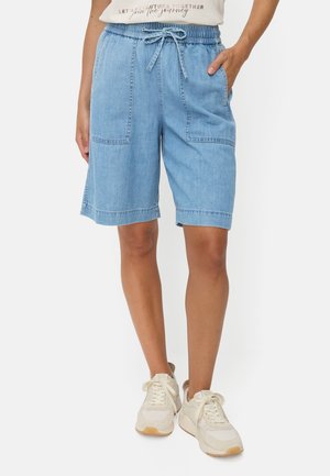 Kvinde iført lyseblå denim knælange shorts med forlommer, snøre i taljen, hvide sneakers og en hvid t-shirt.