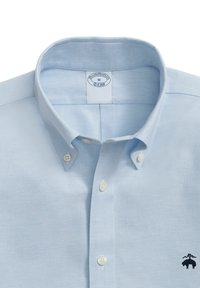 Světle modrá košile na knoflíky z bavlněné tkaniny. Má špičatý límeček, bílé knoflíky a vyšívané logo na levé části hrudi.