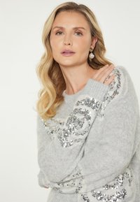 Maglione grigio chiaro e fluffy con pailettes argentate disposte in un motivo circolare lungo le maniche e il corpo, dotato di polsini a coste per una maggiore consistenza.