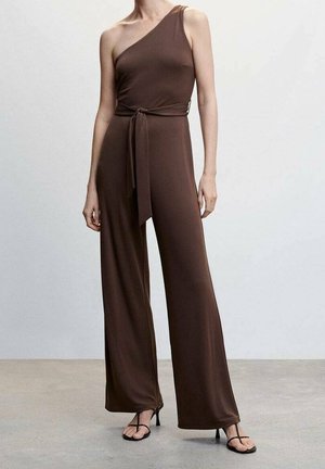 Combinaison marron à une épaule avec ceinture à nouer. Présente des jambes larges et un tissu doux. Le modèle la porte avec des sandales noires à lanières sur un fond neutre.