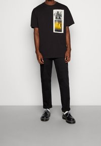 Schwarzes Baumwoll-T-Shirt mit einem grafischen Druck der Buchstaben "CK" und einem gelben Farbverlauf. Getragen mit schwarzen Hosen und schwarzen Schuhen.