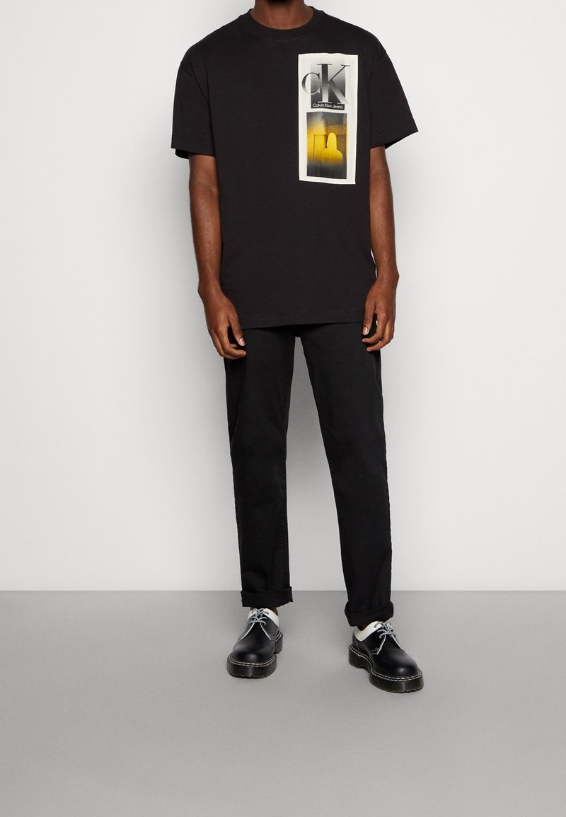 Schwarzes Baumwoll-T-Shirt mit einem grafischen Druck der Buchstaben "CK" und einem gelben Farbverlauf. Getragen mit schwarzen Hosen und schwarzen Schuhen.