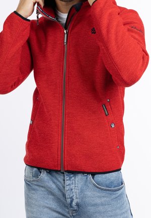 Man draagt een rood jack met rits en zijzakken en borduurwerk op de borst en mouw, gecombineerd met lichtblauwe jeans.