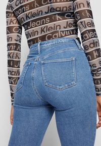 Úzké modré denimové džíny se středním omytím, s zadními kapsami a logem Calvin Klein na pásku. Textura se zdá být hladká.