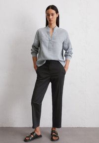 Chemise en lin bleu clair à manches longues avec un col en V sans col, rentrée dans un pantalon noir ajusté, accompagnée de sandales noires avec des accents dorés.