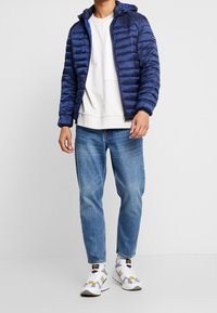 Donkerblauwe pufferjas met capuchon, ritssluiting en doorgestikt ontwerp, gedragen over een lichtgrijze sweatshirt, gecombineerd met lichtblauwe jeans en sneakers.