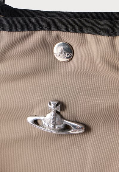 Ασημένιο λογότυπο σφαίρας Vivienne Westwood και κουμπί σε μπεζ ύφασμα με μαύρο περίγραμμα, που δείχνει λεπτομερή υφή και ραφές.