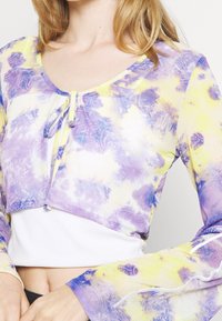 Długi rękaw, krótki top wykonany z przejrzystego materiału w purpurowo-żółty wzór tie-dye. Posiada okrągły dekolt i wiązanie z przodu.