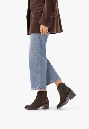 Personne portant un jean court bleu clair, des bottines en daim marron foncé à talons carrés, et une veste en daim marron avec des boutons sur les manches.