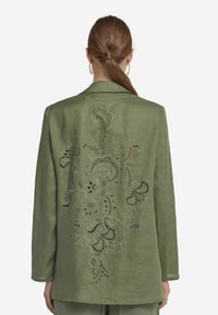 Grön linneblazer med ett utskuret blommönster på ryggen. Har långa ärmar och en klassisk krage. Slät textur överallt.