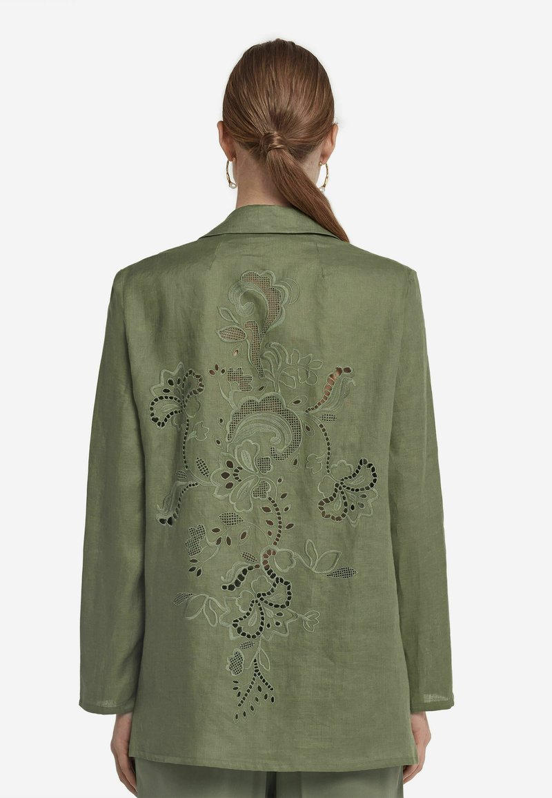 Grön linneblazer med ett utskuret blommönster på ryggen. Har långa ärmar och en klassisk krage. Slät textur överallt.