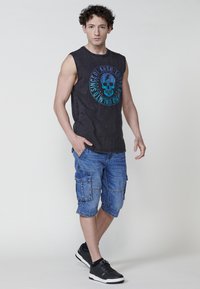 Svart ärmlös tröja med blått dödskallemotiv, denim cargo shorts med sidofickor och svarta träningsskor. Enkel, avslappnad stil.