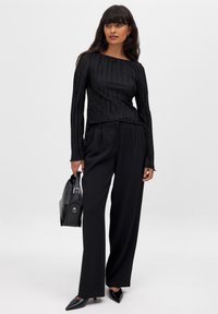 Black pleated long-sleeve top paired with high-waisted black trousers and pointed-toe heels, accented with a structured black handbag.