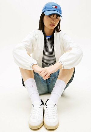 Junge Person sitzt im Schneidersitz, trägt eine blaue Mütze, eine weiße Jacke, ein gestreiftes Hemd, Jeansshorts, weiße Socken und weiße Turnschuhe.