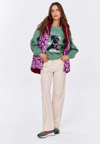 Kobieta w zielonej bluzie z grafiką, z psem, w fioletowej kamizelce w kwiaty, beżowych spodniach i zielono-różowych sneakersach, stojąca z rękami w kieszeniach.