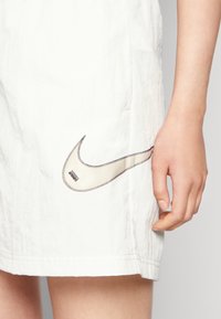White sports shorts made of lightweight material featuring a stitched beige Nike logo on the left side, with visible seams and slight texture.