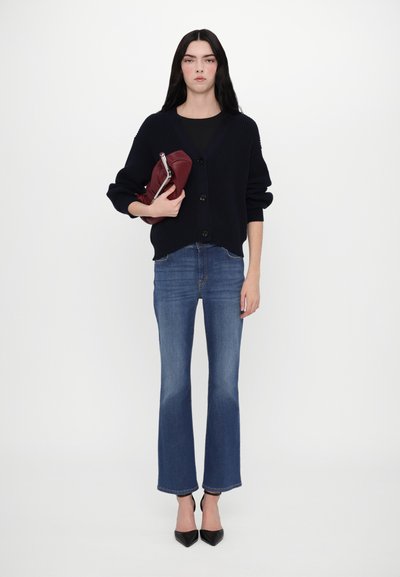 Marineblau gerippter Cardigan, schwarzes Oberteil, hellblaue Schlagjeans und schwarze High Heels. Hält eine bordeauxrote Clutch mit silbernem Beschlag.