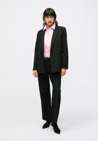 Blazer noir à double boutonnage, chemise rose à col, et pantalon noir sur mesure. Tissu texturé avec des lignes nettes et un design ajusté. Chaussures noires.