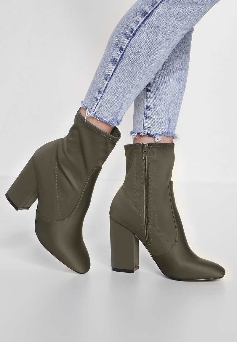 Personne portant des bottines vert olive à talons carrés et fermetures éclair sur les côtés, associées à un jean skinny bleu clair à ourlet effiloché.