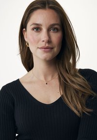 Top nero a righe con maniche lunghe e scollo a V, abbinato a una collana dorata con un piccolo pendente nero. Capelli naturali che scendono sulle spalle.