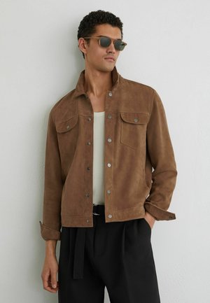 Muž si na sebe obliekol hnedú semišovú bundu na gombíky, biely rebrovaný tielko, čierne nohavice a slnečné okuliare, stojí opretý o bielu stenu.