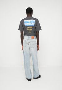 Hombre de espaldas, vistiendo una camiseta gráfica oscura con texto amarillo y un pantalón vaquero claro y holgado, frente a un fondo blanco liso.