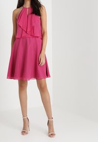 Fuchsia halterneck-klänning med lager av volanger, utställd kjol och nyckelhålsringning. Tillverkad av lätt, flödande tyg.