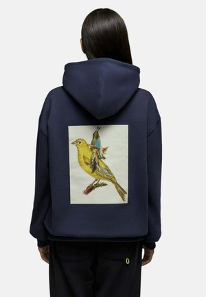 Personne portant un sweat à capuche bleu foncé avec un motif imprimé d'un oiseau jaune et une figure amérindienne assise sur le dos de l'oiseau.